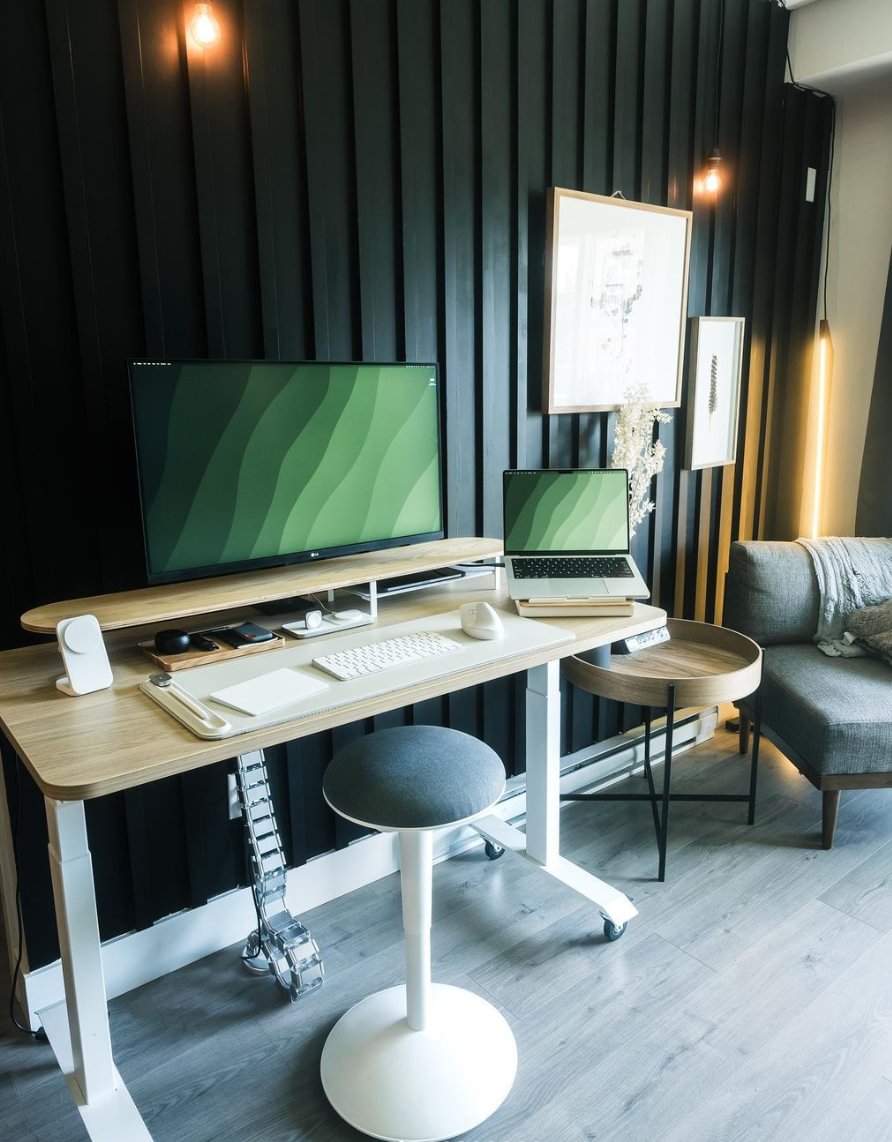A modern desk setup with black wood panelled wall, light wood desk and a white stool