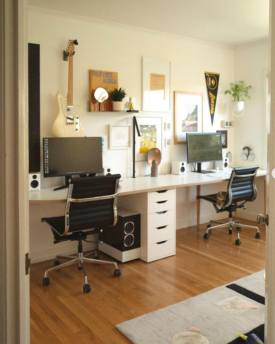 A room with a long desk with 2 computer setups, and a guitar on the wall.