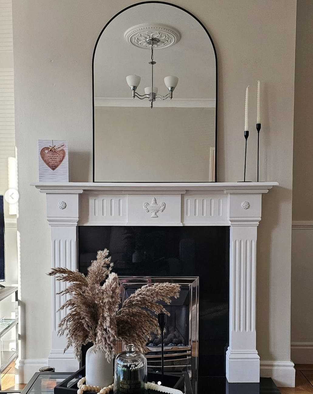 Neutral chimney breast with a white framed fireplace, neutral decorative pieces and a large arched mirror